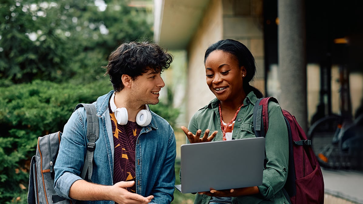 Two students walk around, smile whilst looking at a laptop screen