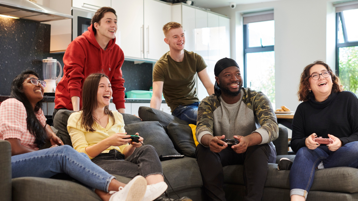 A diverse group of students, sat on a sofa in a communal living area, laughing, talking, and gaming.