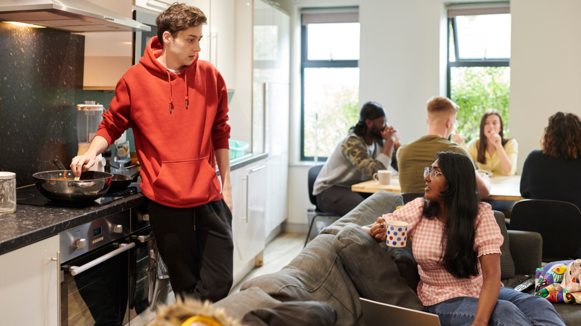 UCAS_Media_ArticleAccom002_Image1 A group of students in a living room, some engaged with a laptop, others cooking, showcasing a casual study space.