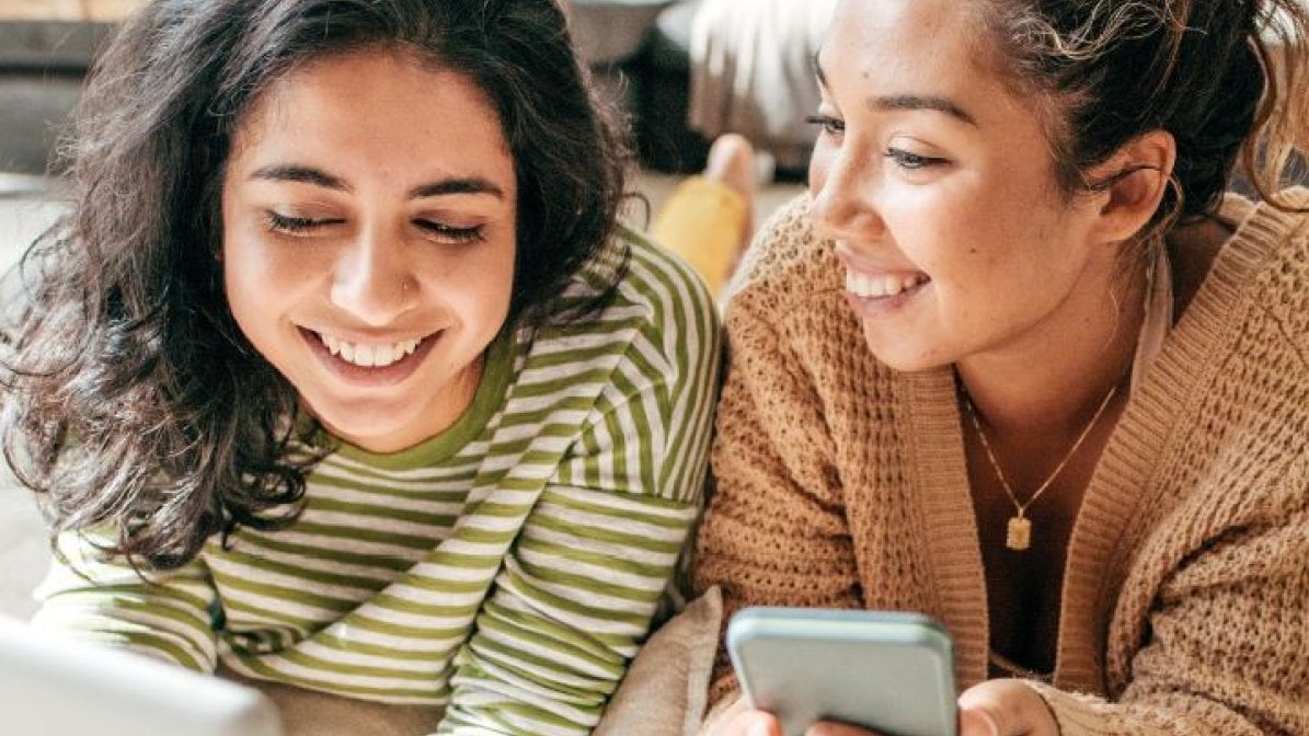 An image of two students, smiling, using a laptop and mobile phone