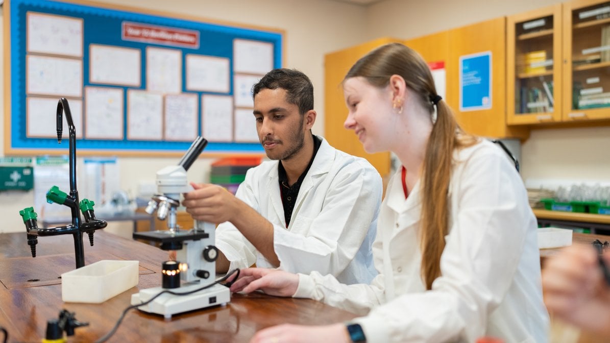 Industry_Marketing_Early_Careers_Article_010324 Two students wearing lab coats sat in a classroom using a microscope