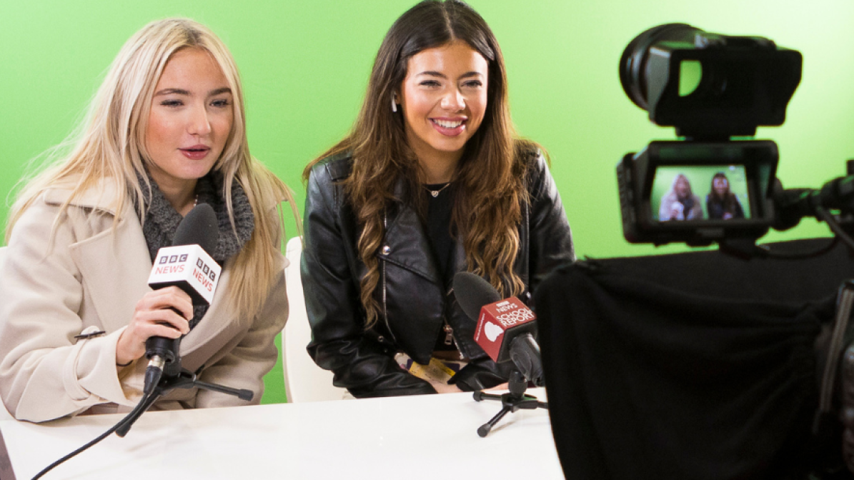 Marketing_Services_Events_Exhibit_As_Employer_Image_Box_1 Two students sat in at a mock news desk, in front of a green screen, with BBC branded microphones in front of them. They're pictured laughing and talking into a studio camera.