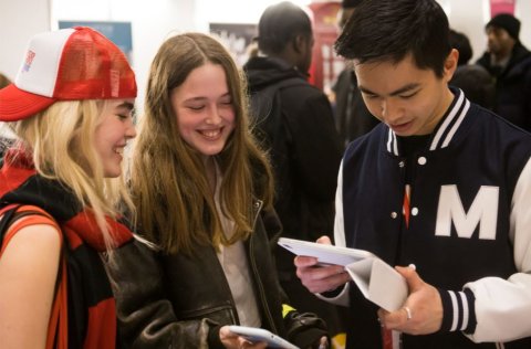A group of three smiling young people are gathered around, looking at a tablet device
