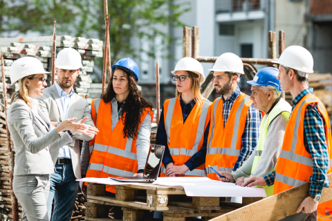 A group of seven constructions workers, in hi vis jacket, stood around a project plan talking