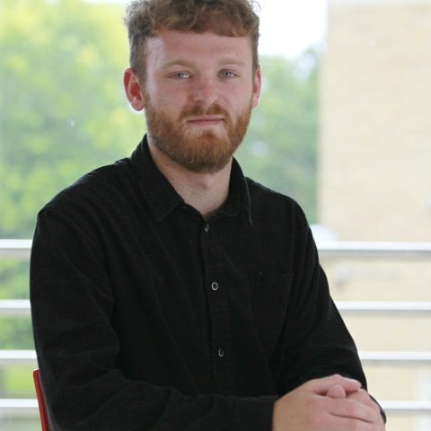 A photo of a young caucasian mail, looking directly at the camera. He's sat down in front of a green, leafy backdrop, with light cascading in from above.